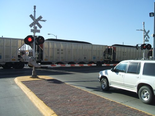 A white SUV waits at a railroad crossing with a train passing by, signaling lights flashing in the background.