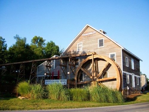 A wooden waterwheel beside a rustic hotel building, surrounded by greenery and a clear blue sky.