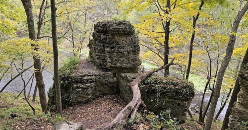 A rocky outcrop surrounded by trees with autumn leaves, overlooking a serene river in a wooded area.