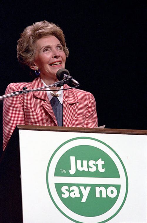 A woman in a pink checkered suit smiles while speaking at a podium with a "Just Say No" sign.