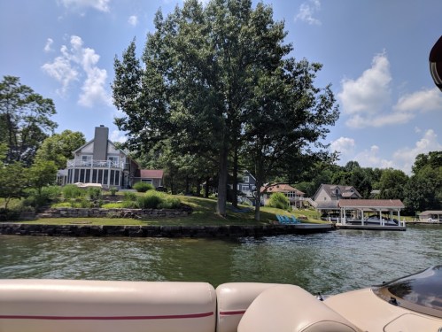 A scenic view of a lake with houses and a large tree on the shore, seen from a boat.