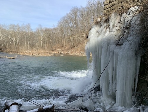Icicles hang from a rocky ledge by a flowing river, surrounded by bare trees and a winter landscape.