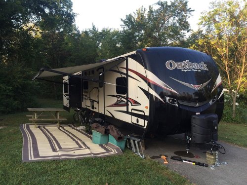 A parked RV with an extended awning, picnic table, and outdoor rug in a wooded campsite setting.