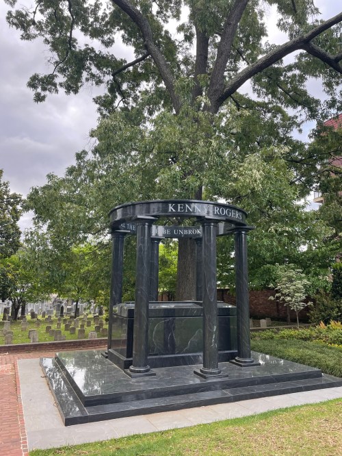 A black marble mausoleum with columns, inscribed with "Kenny Rogers," surrounded by trees and a cemetery.