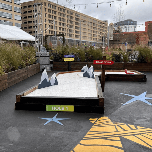 A mini-golf course featuring a snowy mountain theme, with "Hole 1" and "Iceland 2,976 ft" signs. Urban setting in background.