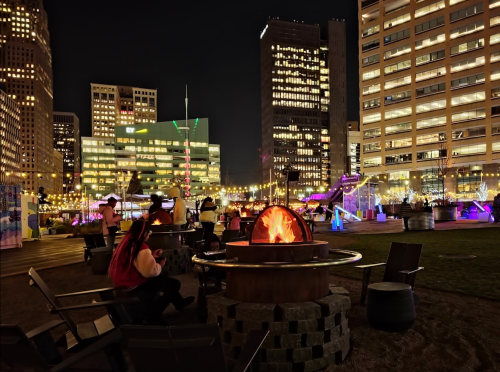 A nighttime scene of a city park with a fire pit, surrounded by people and illuminated buildings in the background.
