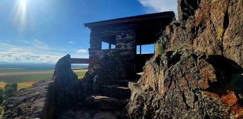 A stone lookout tower perched on rocky terrain, with a scenic view of fields and mountains under a bright blue sky.