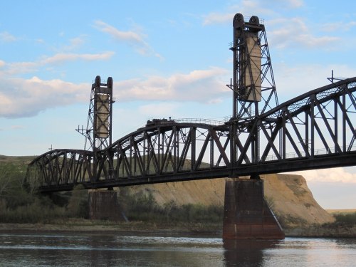 A metal bridge spans a river, featuring tall towers and a curved design against a blue sky with clouds.