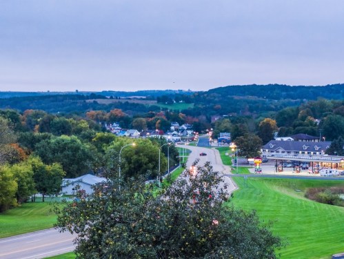 A scenic view of a winding road through a small town, surrounded by trees and hills under a cloudy sky.