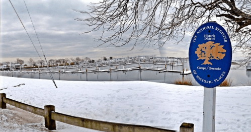 A snowy landscape featuring a calm waterway and a sign for the Historic District of Camp Owasco.
