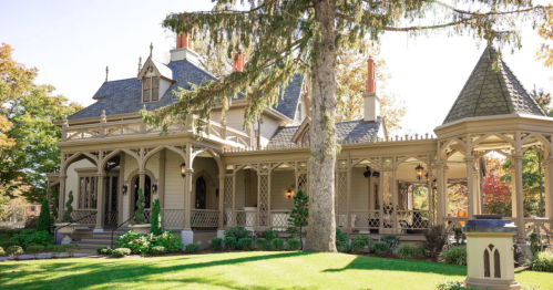 A charming Victorian-style house with a wraparound porch, surrounded by lush greenery and tall trees.