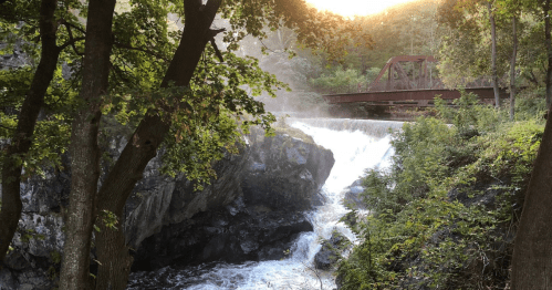 A serene waterfall cascades over rocks, framed by trees, with a red bridge in the background under a glowing sunset.