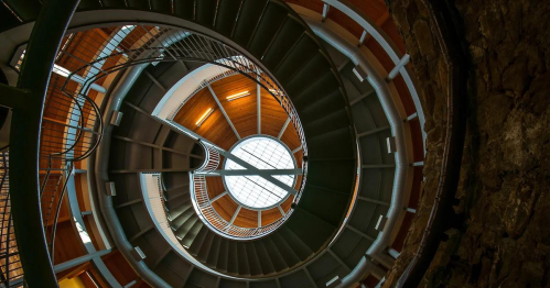 A spiral staircase viewed from below, featuring metal railings and a circular skylight at the top.