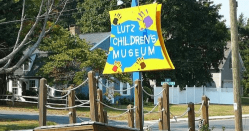 Colorful sign for Lutz Children's Museum with handprints, surrounded by trees and a wooden play structure.