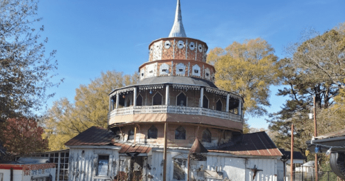 A weathered, multi-tiered building with a spire, surrounded by trees and a clear blue sky.