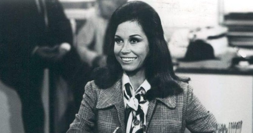A smiling woman in a vintage outfit sits at a desk, with a busy office background.