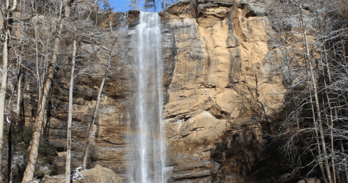 A tall waterfall cascades down a rocky cliff, surrounded by bare trees and a clear blue sky.