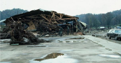 A collapsed building surrounded by debris and muddy ground, with a person inspecting the damage in a rural area.