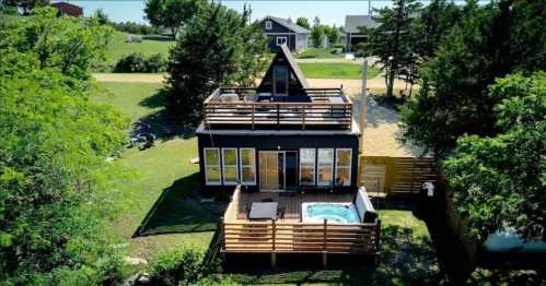 Aerial view of a modern A-frame house with a deck and hot tub, surrounded by greenery and other homes.