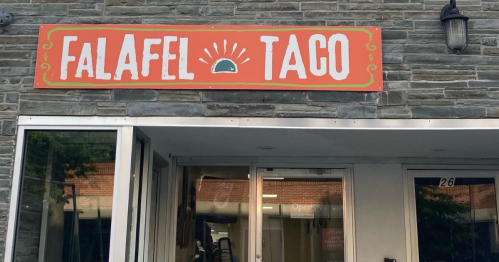 Sign for "Falafel Taco" on a stone wall, featuring a sun and taco graphic. Entrance visible below the sign.
