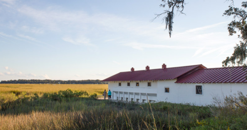 A white building with a red roof beside a grassy field under a clear sky, with a person walking nearby.