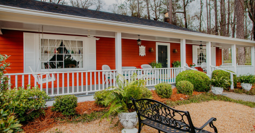 A charming red house with a white porch, surrounded by greenery and decorative plants.