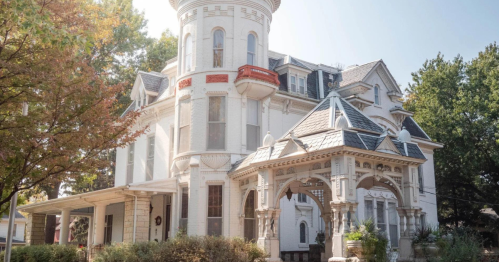 A large, ornate white Victorian house with a turret and decorative porch, surrounded by trees and greenery.