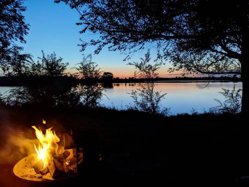 A serene lakeside scene at dusk, featuring a glowing campfire and silhouettes of trees against a colorful sky.