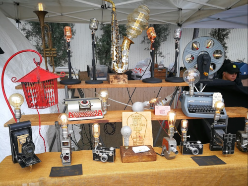 A display of vintage cameras, typewriters, and unique lamps at a market stall, with a saxophone in the background.