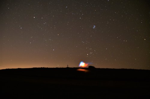 A starry night sky with a colorful glow on the horizon, silhouetting a distant hill against the dark backdrop.