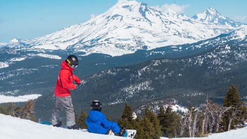 Two snowboarders in winter gear enjoy a scenic view of snow-covered mountains and trees.