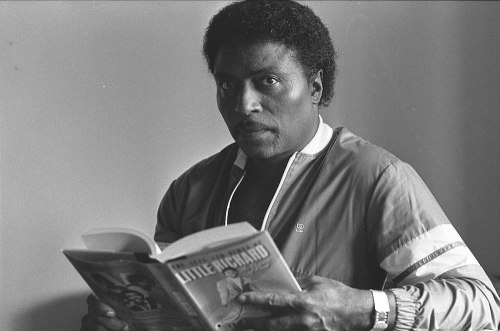 A man in a tracksuit holds a book titled "Little Richard," looking thoughtfully at the camera.