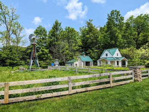 A quaint farmhouse with a green roof, surrounded by trees and a windmill, set in a lush green landscape.