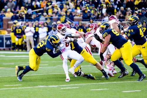 Two Michigan football players tackle an opposing player during a game, with a crowd in the background.
