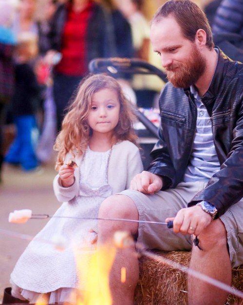 A man and a young girl sit by a campfire, roasting marshmallows, with a warm, festive atmosphere around them.
