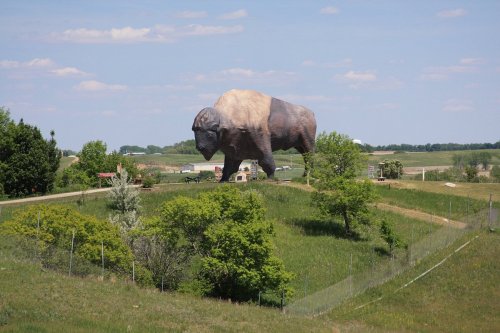 A giant buffalo statue stands in a green landscape with trees and a blue sky in the background.