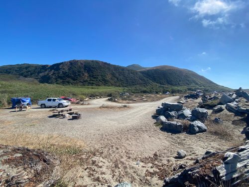 A gravel road leads through a campsite with parked vehicles, surrounded by hills and rocky terrain under a clear blue sky.