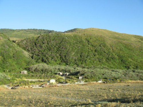 A green hillside with small structures at its base, under a clear blue sky.