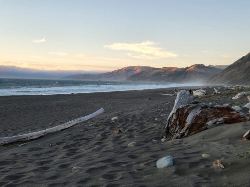 A serene beach scene at sunset, featuring gentle waves, sandy shore, and distant mountains under a colorful sky.