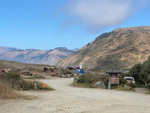 A dirt path leads through a scenic landscape with hills, a few vehicles, and a signpost in the foreground.