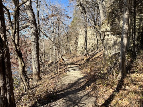 A winding dirt path through a wooded area with rocky cliffs and bare trees under a clear blue sky.