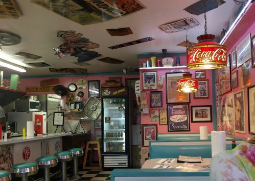 A retro diner interior with colorful decor, vintage signs, and a checkered floor, featuring teal booths and a Coca-Cola lamp.