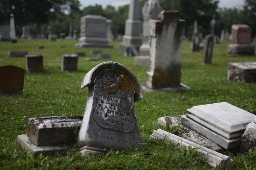 A weathered gravestone lies tilted in a grassy cemetery, surrounded by other gravestones and memorials.