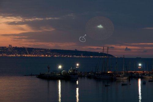 A twilight scene of a harbor with boats, illuminated by lights, and a mysterious cloud in the sky above the coastline.