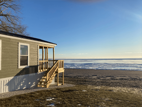 A beach house with a wooden deck overlooks a sandy beach and calm water under a clear blue sky.