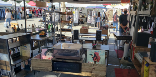 A vibrant market stall featuring vintage records, speakers, and various bottles, with shoppers browsing in the background.