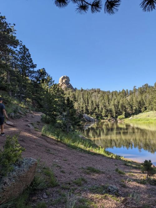 A serene lakeside scene with trees, rocky formations, and a clear blue sky in the background.