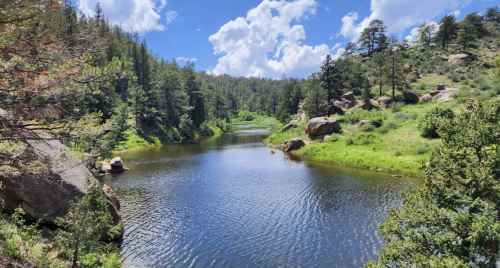 A serene river flows through a lush green landscape, surrounded by trees and rocky hills under a blue sky with clouds.