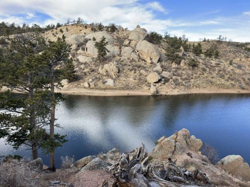 A serene lake surrounded by rocky hills and pine trees under a blue sky with scattered clouds.
