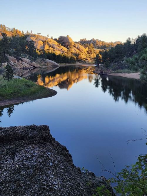 A serene lake reflects rocky hills and trees at sunset, surrounded by lush greenery.